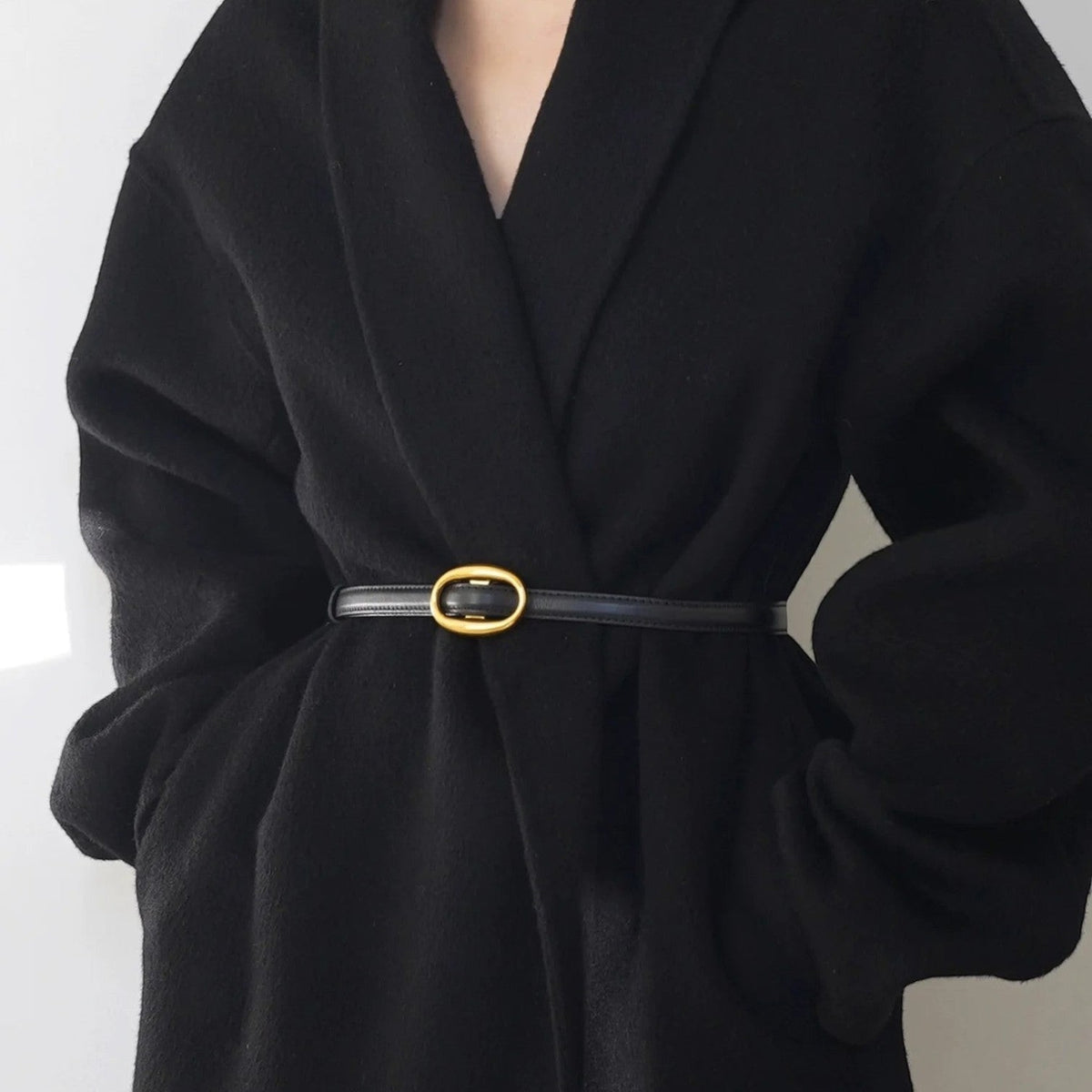 Ceinture Ceintura Capri fine en cuir noir avec boucle ovale dorée portée sur un manteau noir élégant